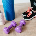 Unrecognizable sportswoman wearing sneakers and black leggings standing with sport mat on wooden floor near dumbbells and water bottle before exercising indoors
